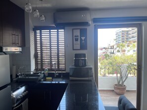 Una cocina moderna con una isla central, estufa, fregadero y cafetera. Hay una ventana con persianas, una planta en maceta en el balcón y vistas a un área residencial.