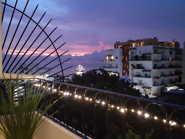 Un balcón con luces de cuerda, con vistas a una zona costera con edificios y una isla distante.