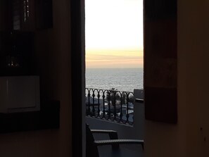 A room with a view of the sea at sunset, a balcony with a railing, and a chair.