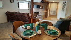 A dining area with a round table set for four, surrounded by chairs. A brick fireplace is visible in the background, and a television is mounted on the wall above it.