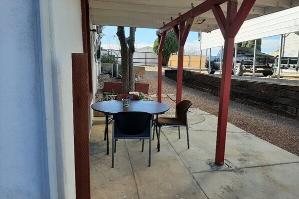 An outdoor patio with a table and chairs, a covered area with a red beam structure, and a view of parked vehicles and a building in the background.
