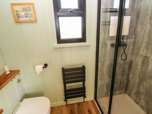 A bathroom with a toilet, a towel rack, a shower with a glass door, and a small window. - The Honeybee (Pembroke)