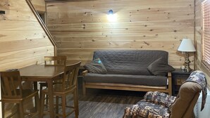 A cozy cabin interior with a wooden ceiling, a sofa, a dining table with chairs, and a patterned throw blanket on a chair.