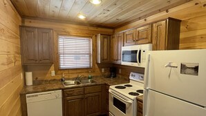 A compact kitchen with wooden walls and ceiling, a white dishwasher, stove, and refrigerator, and a window with blinds.