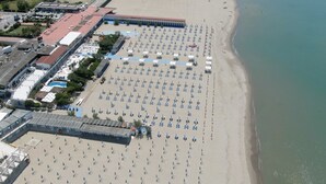 An aerial view of a beach with rows of sun loungers and umbrellas, a building with a red roof, and a clear view of the ocean.