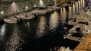 A marina at dusk with boats docked along a waterfront, a stone wall, and buildings in the background.