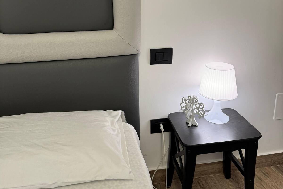 A neatly made bed with a grey headboard, a bedside table with a lamp, and a power outlet.