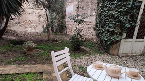 Un tavolo bianco all'aperto con teiera, tazze e una sedia, immerso in un giardino con muri in pietra e vegetazione.
