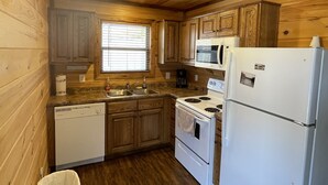 A compact kitchen with wooden walls and ceiling, a dishwasher, a stove, a refrigerator, and a window with blinds.