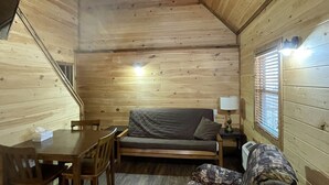 A cozy cabin interior with a wooden ceiling, a sofa, a table with chairs, and a patterned blanket on the floor.