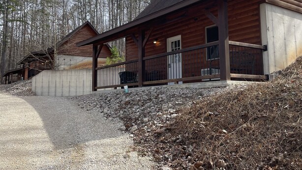 A wooden cabin with a porch, a gravel driveway, and a forested backdrop.