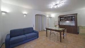 A living room with a blue sofa, a dining table with chairs, a kitchen area with cabinets, and a ceiling light fixture. - House "Le Anfore" - Amalfi Coast (Conca dei Marini)