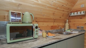 Eine kompakte Küche mit einer Vintage-Mikrowelle in Grün, einem Wasserkocher und einem Toaster. Es gibt ein Spülbecken mit einem Messerblock und ein Teller mit einem Croissant.