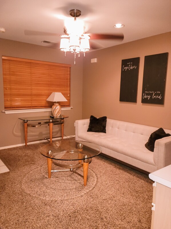 A living room with a white sofa, a glass coffee table, and two side tables. There is a window with blinds, a chandelier, and two wall art pieces.