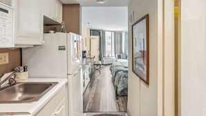 Un pasillo estrecho con una cocina a un lado y un dormitorio al otro. La cocina tiene un fregadero, armarios y un refrigerador. El dormitorio tiene una cama, un ventanal y una puerta que da a otra habitación.