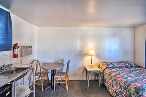 Una habitación compacta con kitchenette, una cama con ropa de cama estampada, una mesita con lámpara y una silla.