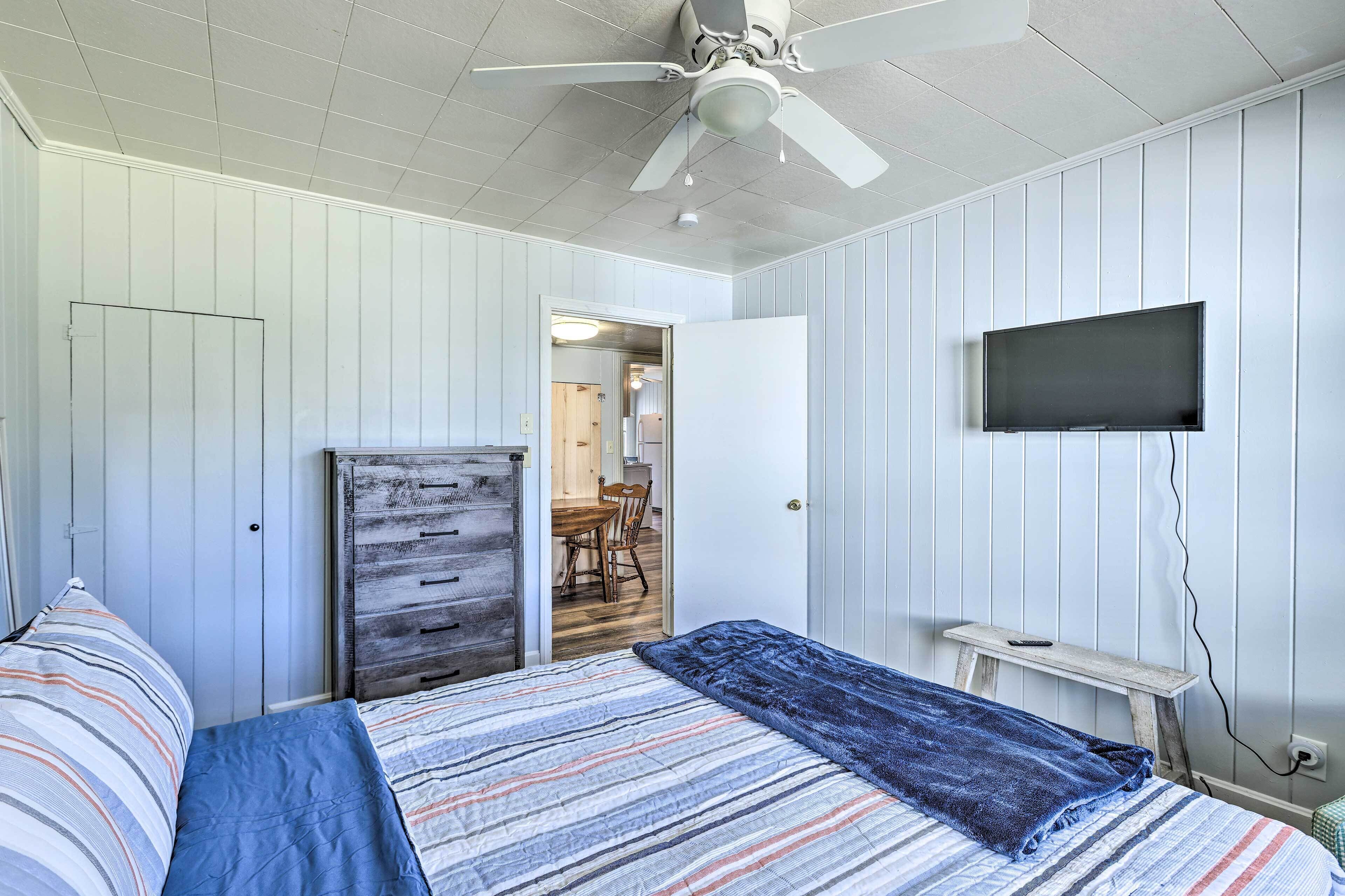 Ein Schlafzimmer mit einem Bett, einem Deckenventilator, einem an der Wand montierten Fernseher und einer hölzernen Kommode.