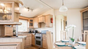 A compact kitchen with wooden cabinets, a stove, and a dining area with a table set for two. - Mountain View Lodge 2 (Holyhead)