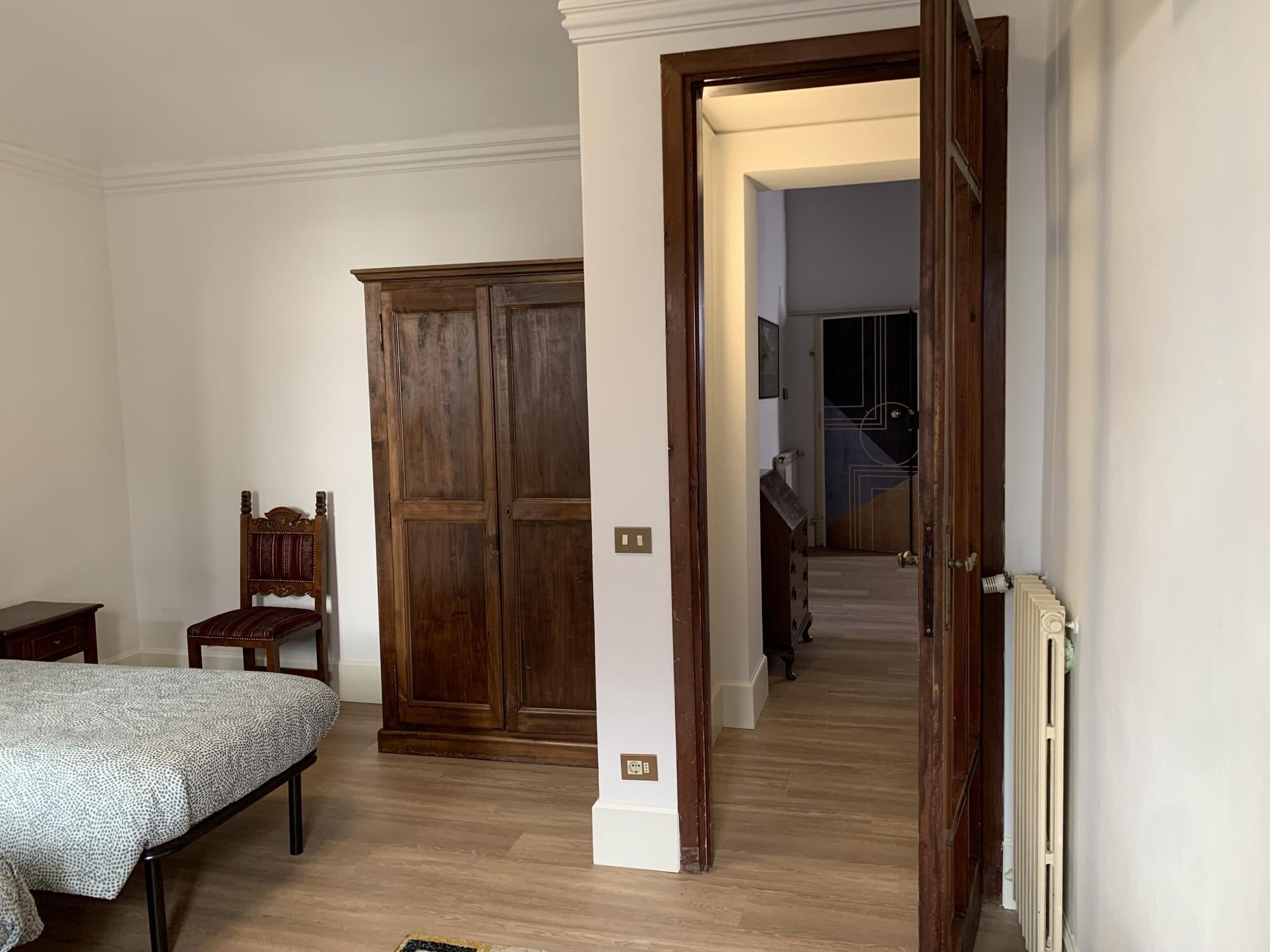 A bedroom with a bed, a wooden wardrobe, and a chair. There is a hallway leading to a staircase.