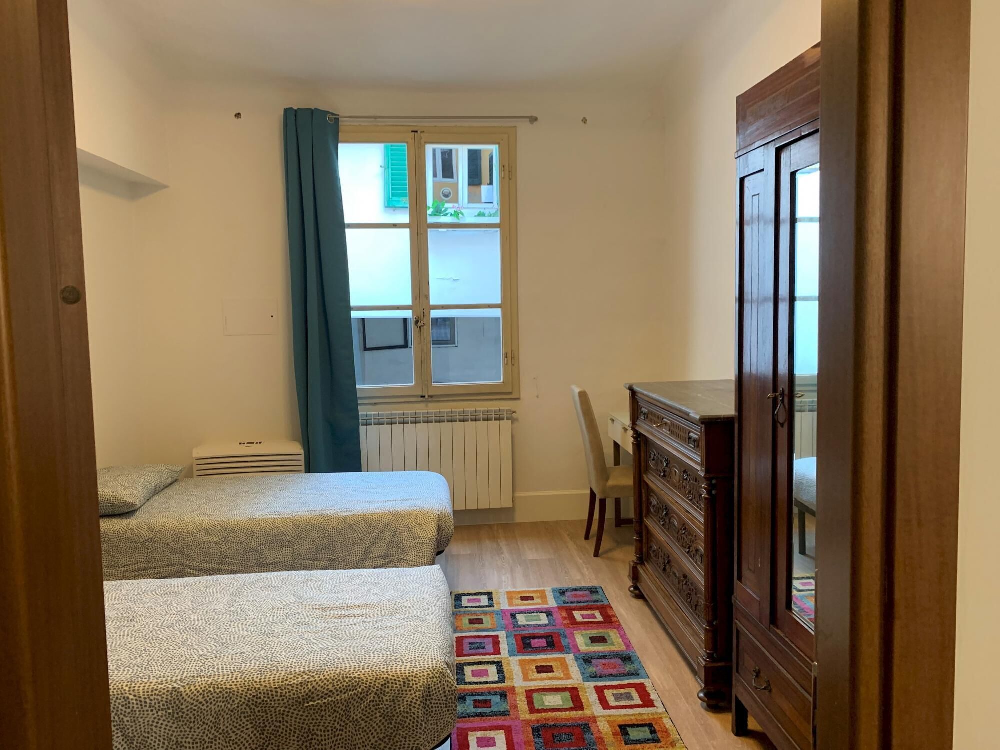 Een kleine, smalle kamer met twee bedden, een radiator, een raam met gordijnen, een houten kast en een kleurrijk vloerkleed.