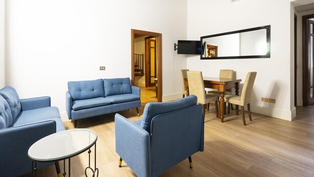 Una sala de estar con un sofá azul, una mesa de comedor con sillas, un televisor de pantalla plana en la pared y piso de madera.