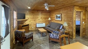 A cozy cabin interior with wooden walls, a ceiling fan, a sofa, a coffee table, and a small table with a lamp.