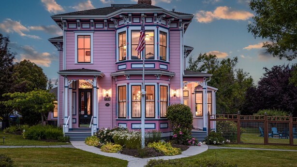 Une maison de style victorien, peinte en rose, avec un porche à l’avant, de grandes fenêtres et un drapeau américain.
