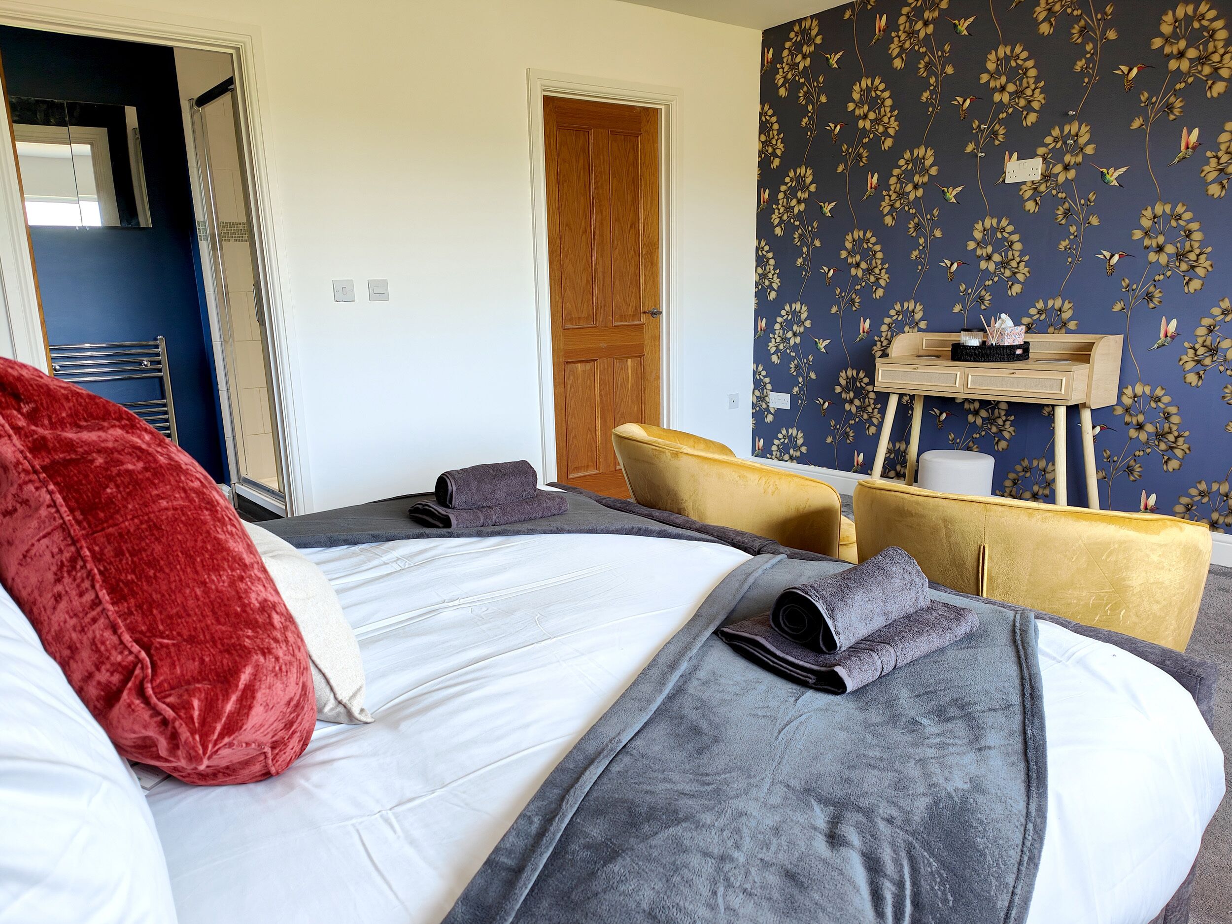 A hotel room with a bed, a wooden door, a mirror, a vanity with a mirror, and a floral wallpaper.