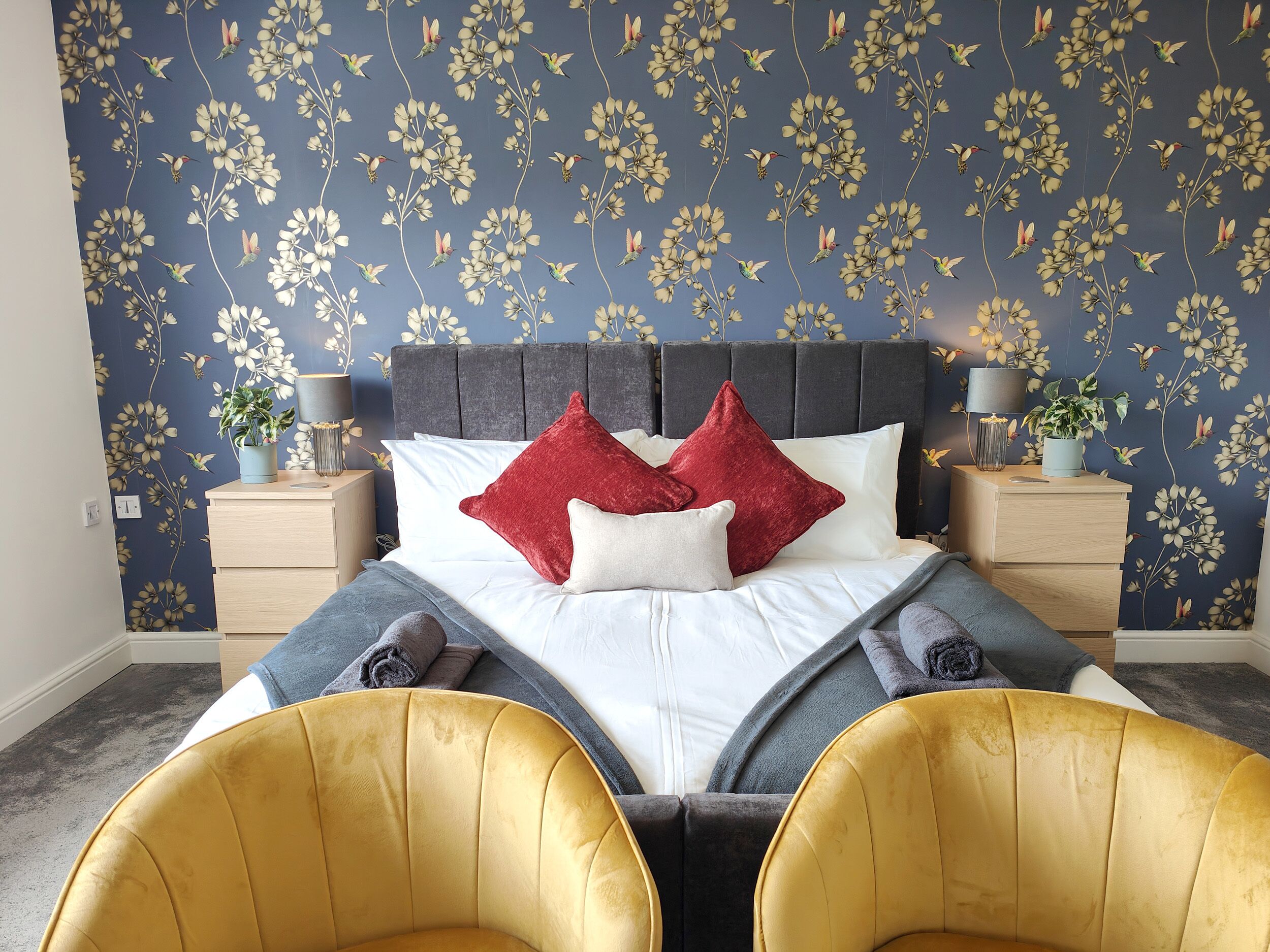 Une chambre avec un papier peint à motifs floraux, un lit avec des coussins blancs et rouges, deux tables de chevet avec des lampes et deux chaises jaunes.