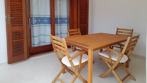 Een houten eettafel met vier stoelen, een schuifdeur met houten luiken en een vloer van witte tegels.