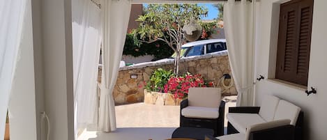 Quarto com mesa de jantar, cadeiras, sofá e vista para um jardim com árvore e flores.