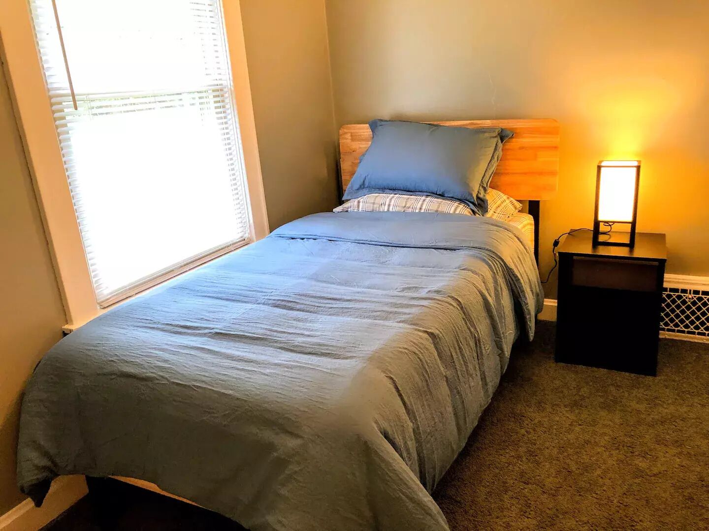 Una cama pequeña y bien hecha con colcha y almohadas azules, una lámpara de noche y una ventana con persianas.