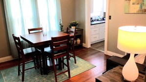 A dining area with a wooden table and chairs, a rug, a lamp, and a kitchen in the background.