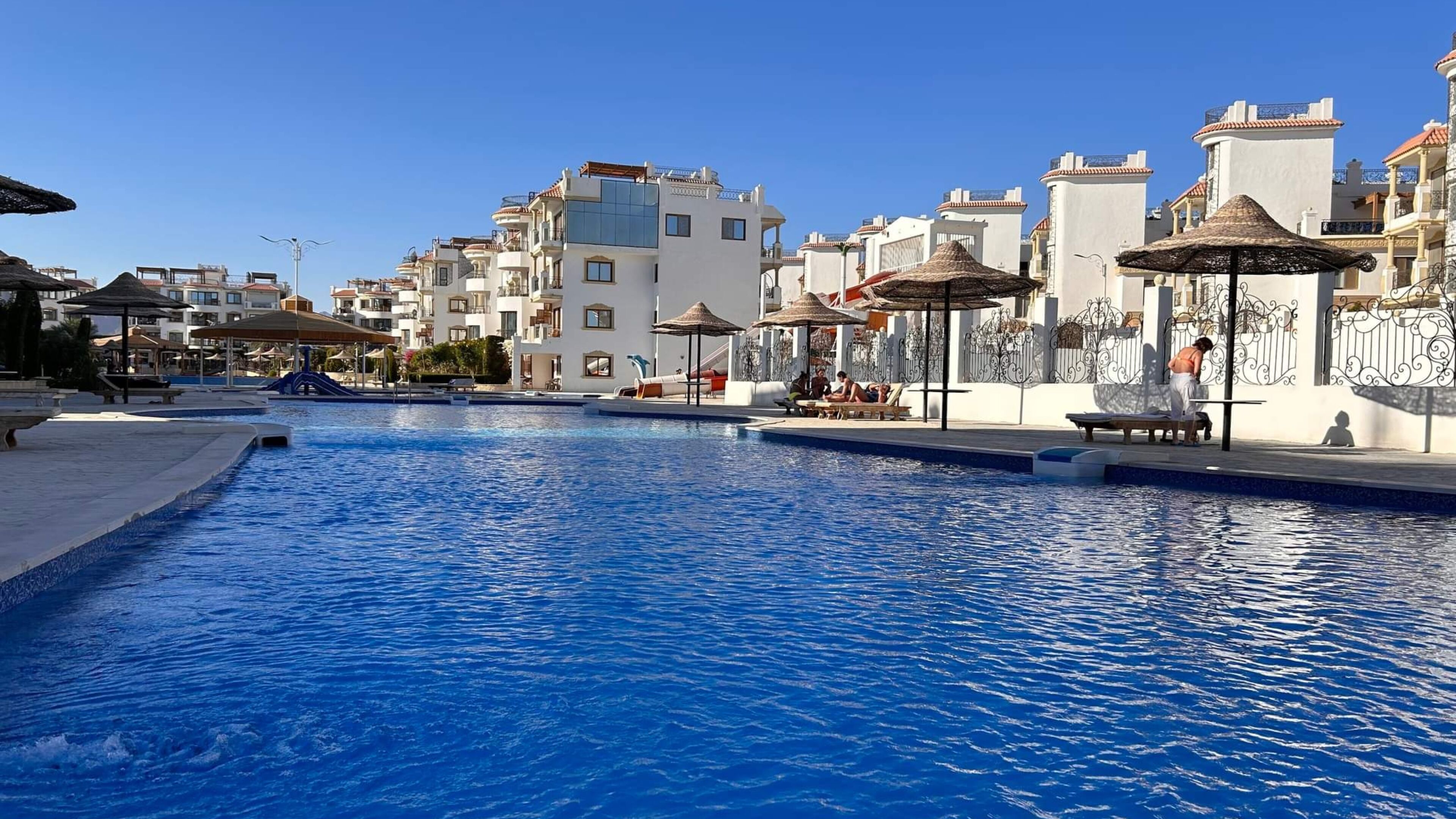 A swimming pool with lounge areas and umbrellas, surrounded by multi-story buildings with balconies.