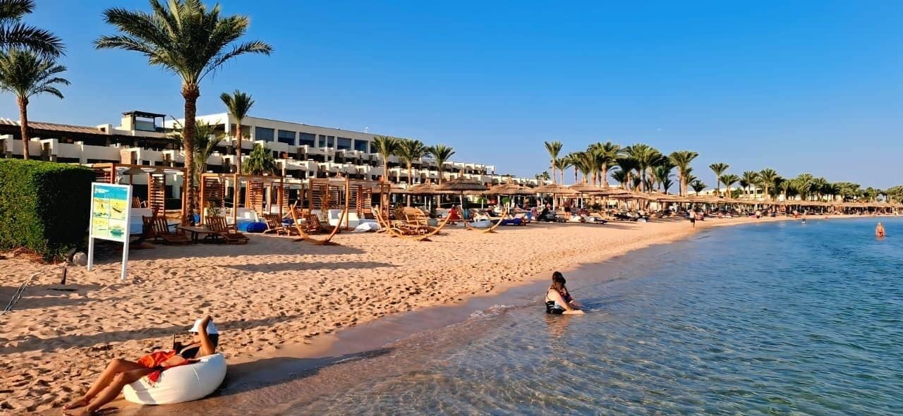 Un complejo turístico en la playa con palmeras, tumbonas y un mar azul cristalino.