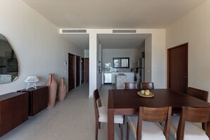 A modern dining area with a wooden table and chairs, a kitchen in the background, and a hallway leading to a door.