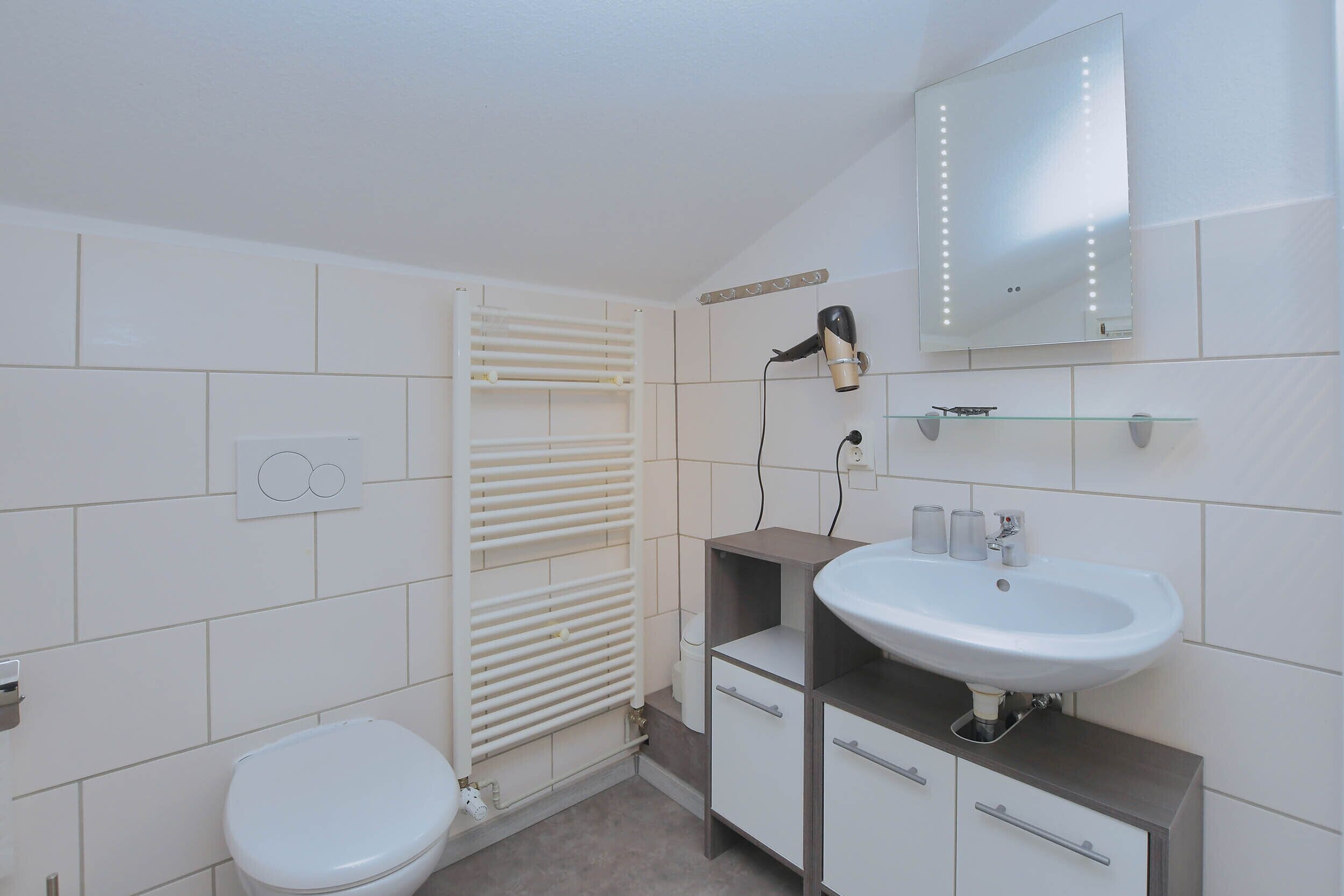 Une salle de bain moderne avec des toilettes blanches, une étagère en verre au-dessus du lavabo, un sèche-cheveux et un miroir avec éclairage intégré.