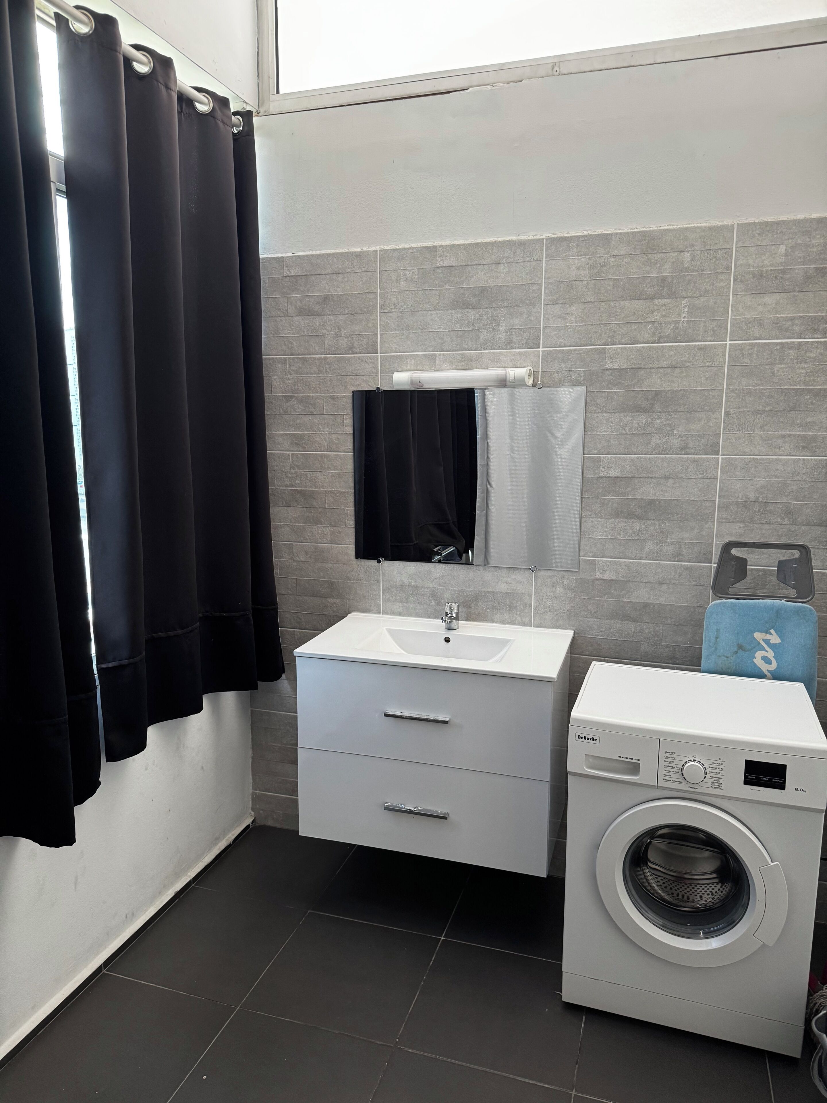 Een badkamer met een witte wastafel, een spiegel, een zwarte handdoek, een wasmachine en grijze betegelde wanden.