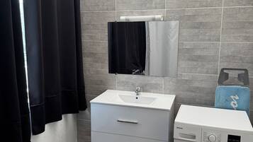 Een badkamer met een witte wastafel, een spiegel, een zwarte handdoek, een wasmachine en grijze betegelde wanden.