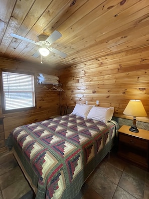A wooden cabin bedroom with a bed, a hanging light fixture, a window with blinds, and a nightstand with a lamp.