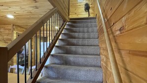 Un escalier en bois, doté d’un palier recouvert de moquette et d’une rampe, mène à une pièce où est installé un ventilateur de plafond.