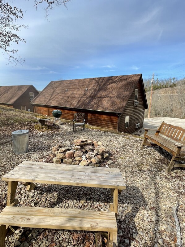 A rustic outdoor area with a wooden bench, a stone fire pit, and a wooden building with a sloped roof.