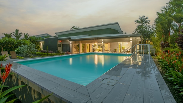 A modern pool area with a clear blue pool, a wooden deck, and a building with large windows and a covered patio. - Villa Margarita - Families Getaway in La Fortuna (La Fortuna)