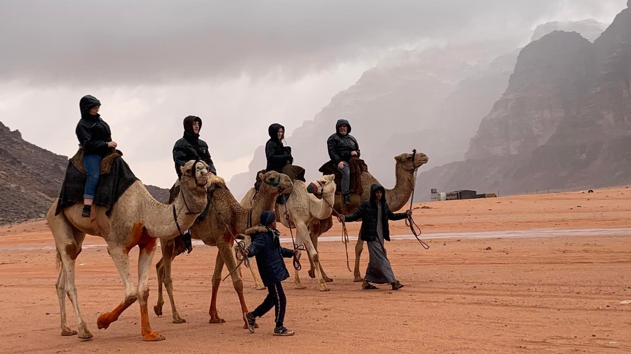 A group of people riding camels in a desert landscape with rocky mountains in the background.