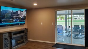Una sala de estar con una televisión de pantalla plana montada en la pared, un mueble de madera para entretenimiento y un patio exterior accesible por una puerta corrediza de vidrio, con una mesa y sillas.