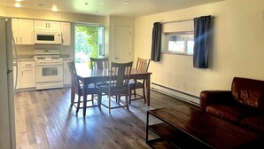 Una cocina compacta con muebles blancos, microondas y estufa. Hay una mesa de comedor con sillas, y un sofá de cuero marrón en una sala de estar con ventana.