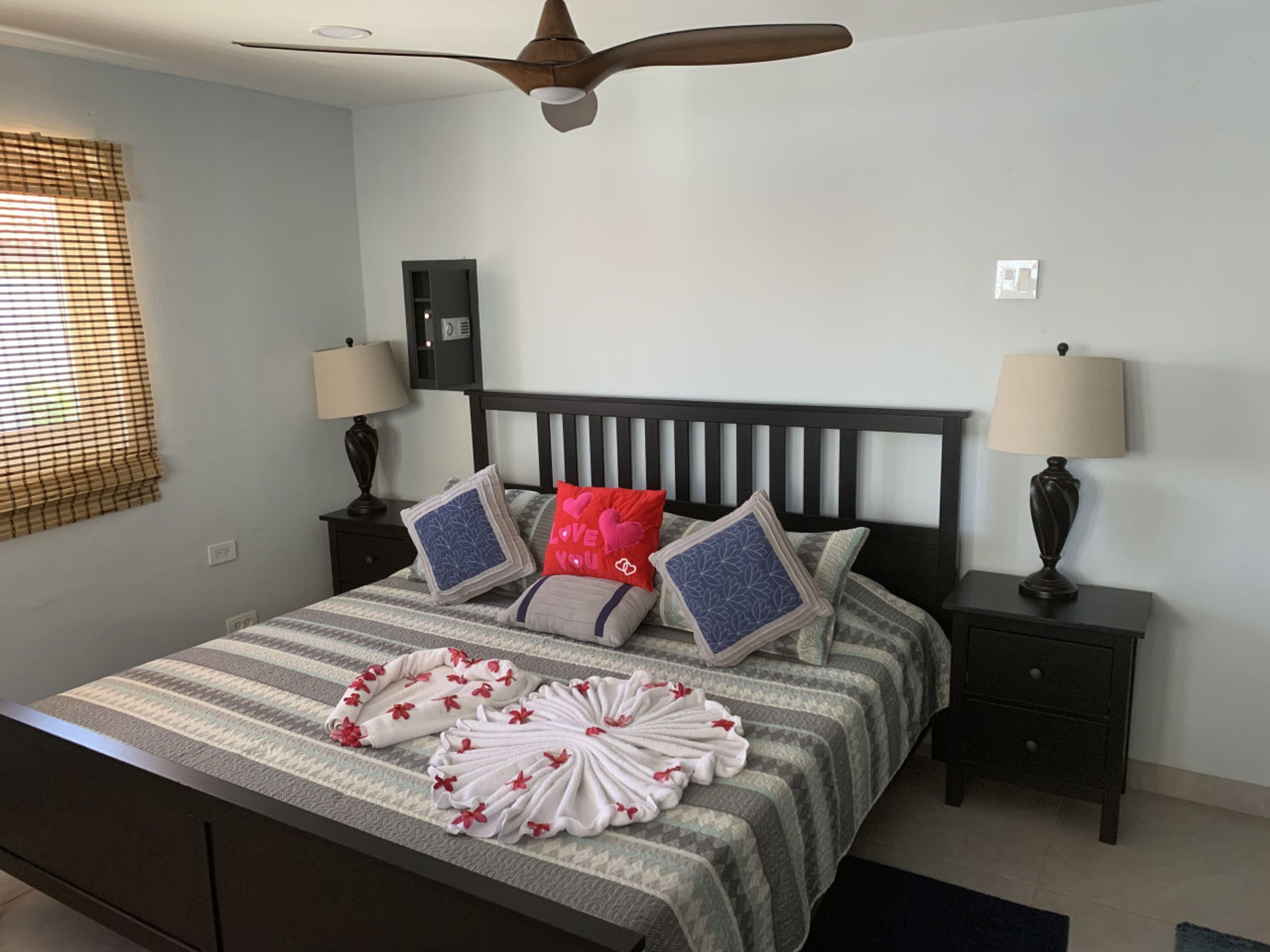 A neatly made bed with striped bedding, two bedside tables with lamps, a ceiling fan, and a window with blinds.