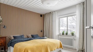 Ein Schlafzimmer mit einem Bett, einem Heizkörper, einem Fenster mit Vorhängen und einer Wand mit vertikalen Linien.