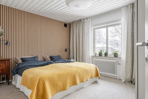 A bedroom with a bed, a radiator, a window with curtains, and a wall with vertical lines.