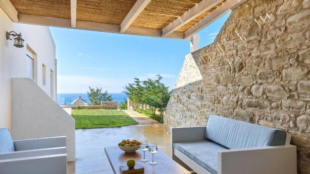 A covered outdoor seating area with a stone wall, a wooden table with a bowl of fruit and glasses, and a view of the sea and a grassy area. - Elkysti Crete Superior Apartment "Nikos" (Kreta)